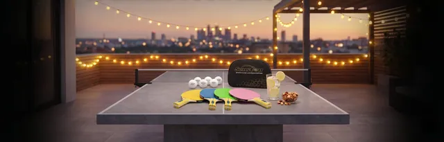 Table tennis paddles, balls and a drink on a rooftop table under string lights at dusk