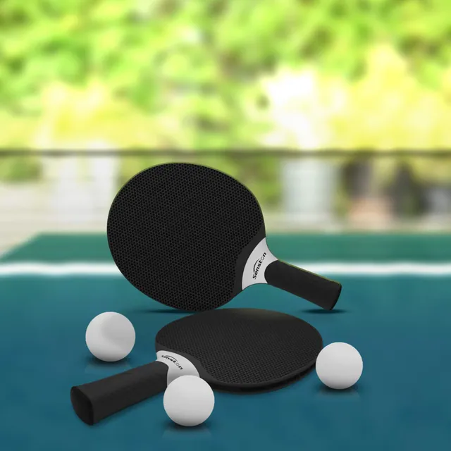 Two black paddles and three white balls on a teal outdoor table with a garden behind