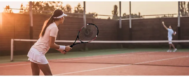 A woman in white waiting for a serve on a clay court at sunset
